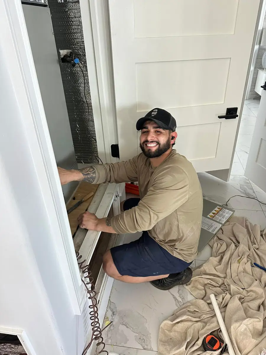 A man kneeling and installing an HVAC air handler in a home utility closet, smiling and wearing a baseball cap.