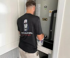 Man servicing a residential HVAC unit in a utility closet, holding a bucket while inspecting components.