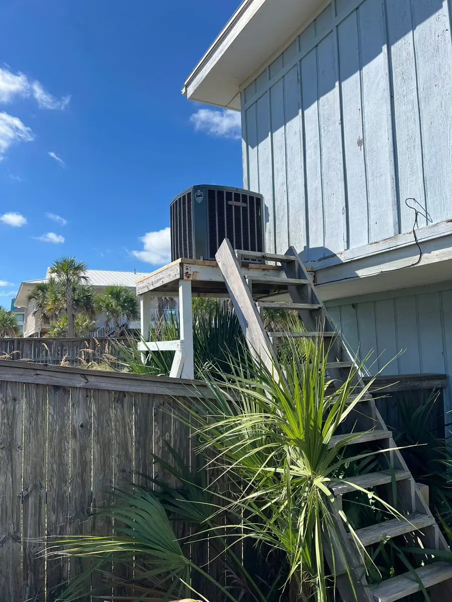 Outdoor air conditioning unit on an elevated wooden platform with stairs, palm plants, fence and blue sky by raised house