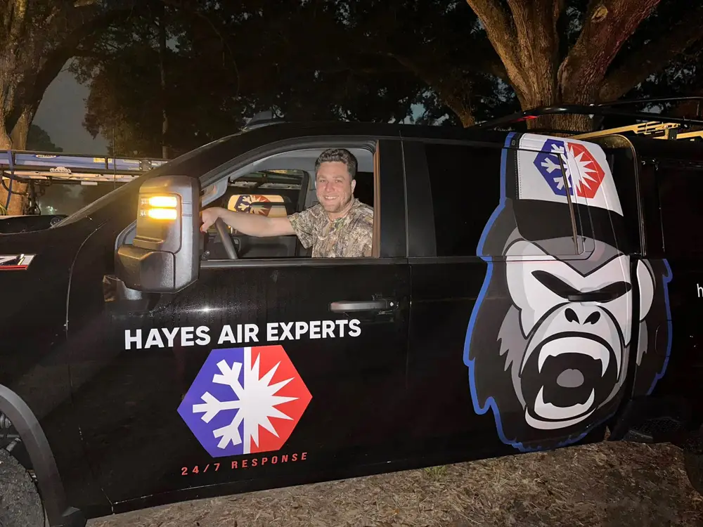A man smiling in driver's seat of a Hayes Air Experts service truck with gorilla logo at night.