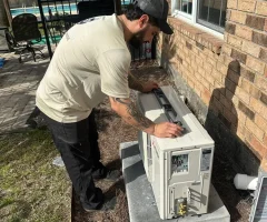 A man installing an outdoor mini-split HVAC unit on a concrete pad beside a brick house