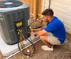 Technician in blue shirt crouching while checking air conditioner with HVAC manifold gauges and colored hoses outdoors.