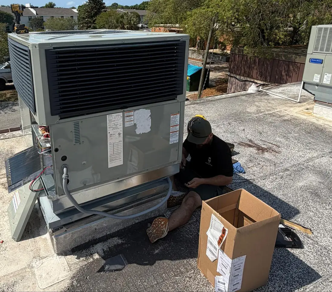 A man working on a rooftop HVAC unit beside an open cardboard box on a flat roof