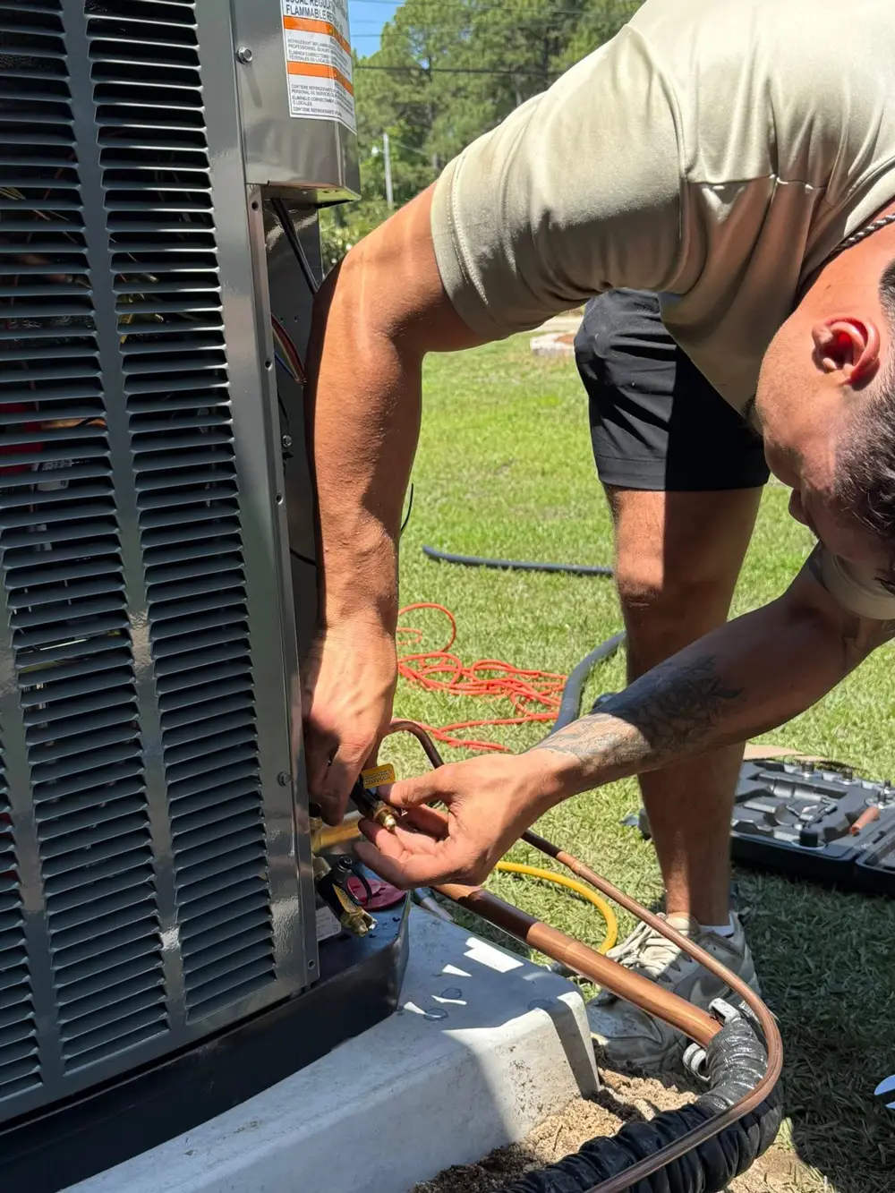 A man servicing an outdoor air conditioner, connecting copper refrigerant lines with gauges and tools on grass
