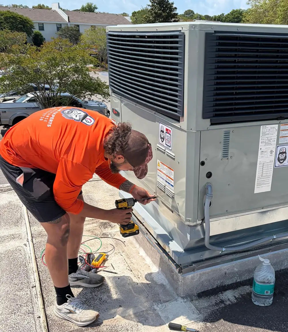 A technician in an orange shirt uses a drill to service a rooftop HVAC unit