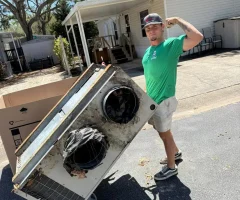 A man in a green shirt flexing while moving a large old HVAC unit on a dolly in a mobile home driveway