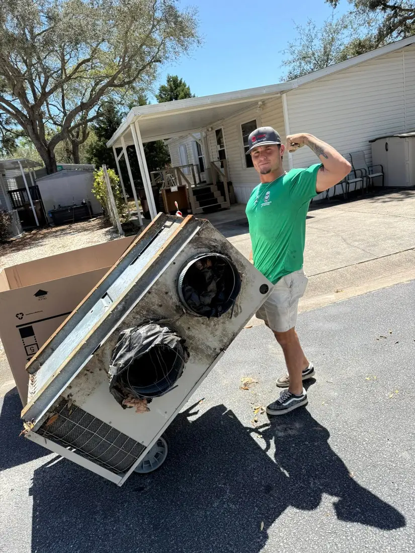 A man in a green shirt flexing while moving a large old HVAC unit on a dolly in a mobile home driveway