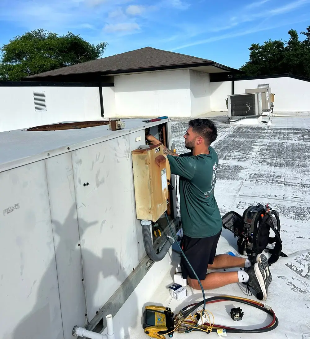 A man HVAC technician kneeling on a rooftop repairing a commercial rooftop air conditioning unit with tools nearby.