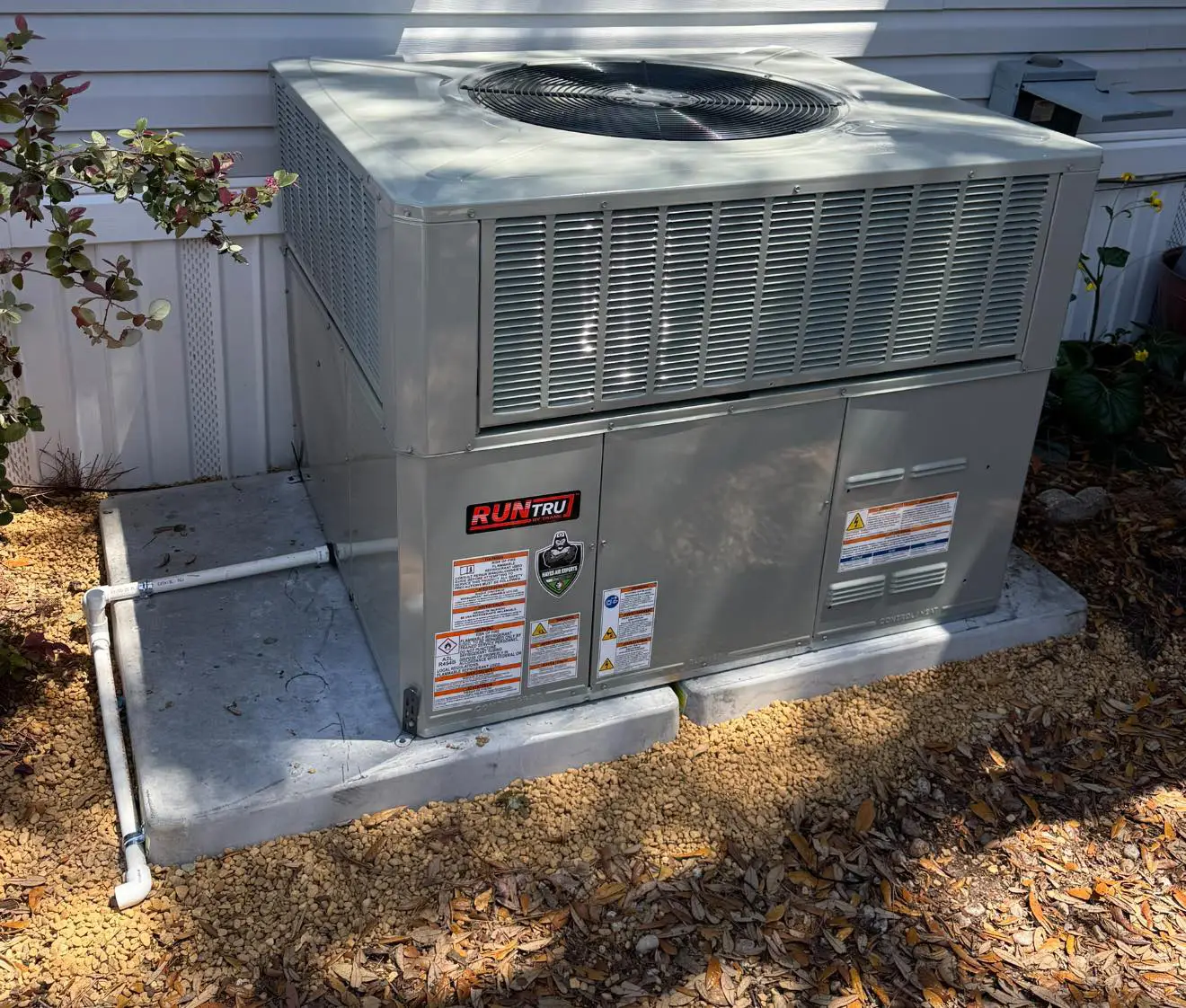RunTru packaged air conditioning unit on concrete pad beside vinyl-sided house, surrounded by gravel and leaves