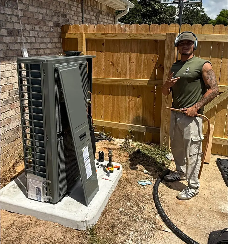 HVAC technician (a man) installing outdoor AC unit on concrete pad, holding copper tubing with tools nearby.