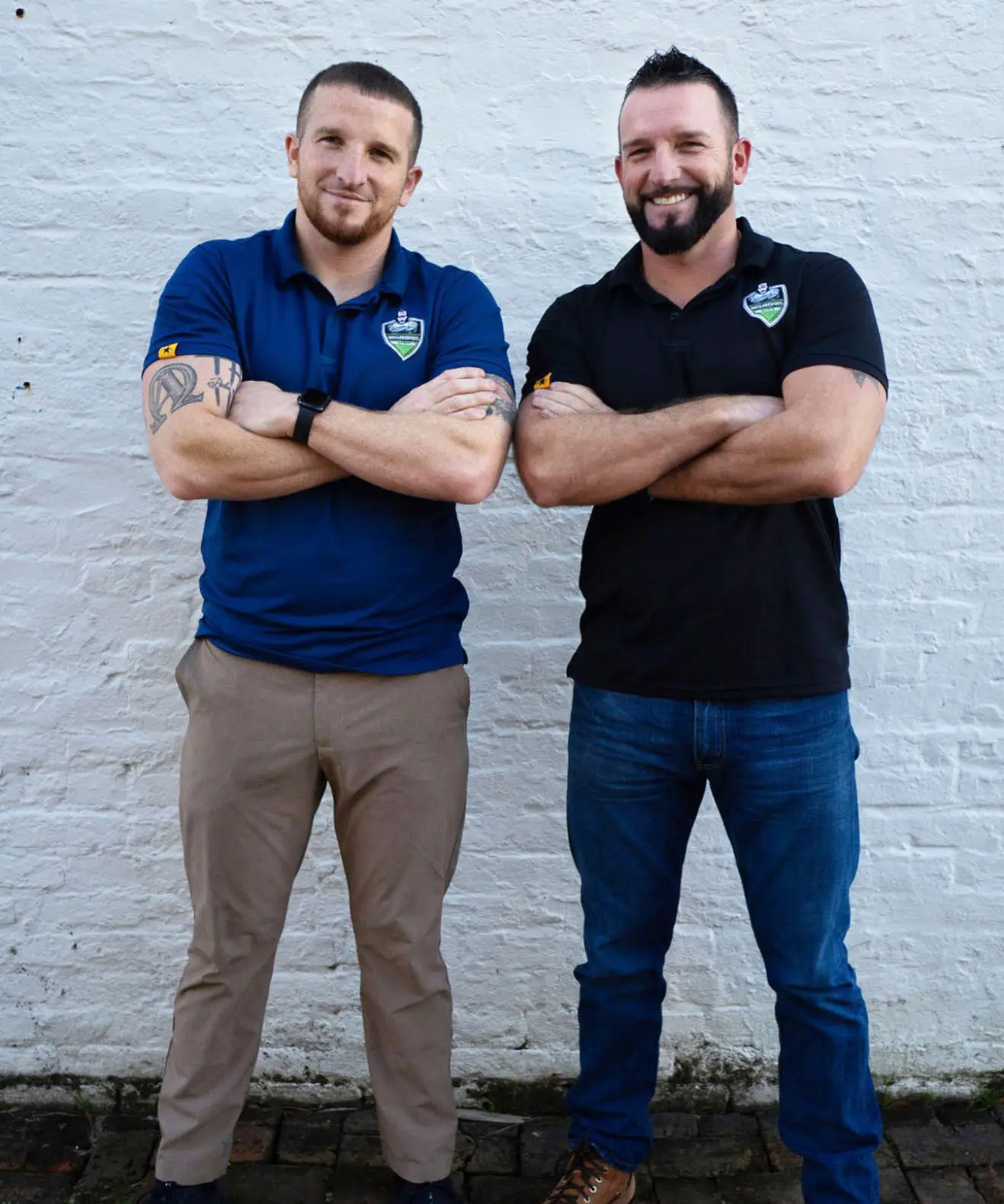 Two men standing side by side against a white brick wall with arms crossed, wearing polo shirts and jeans.