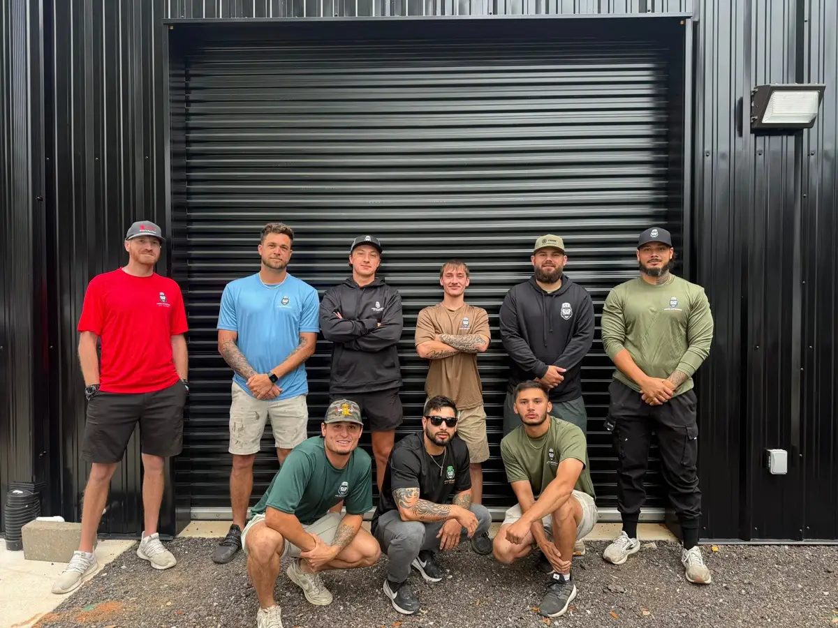 Nine workers posing in front of a black metal building with a closed roll-up door.