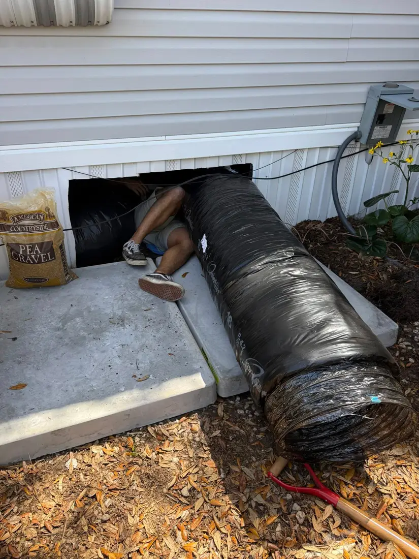 A person crawling into a mobile home crawlspace next to a large black flexible drainage pipe and a bag of pea gravel.