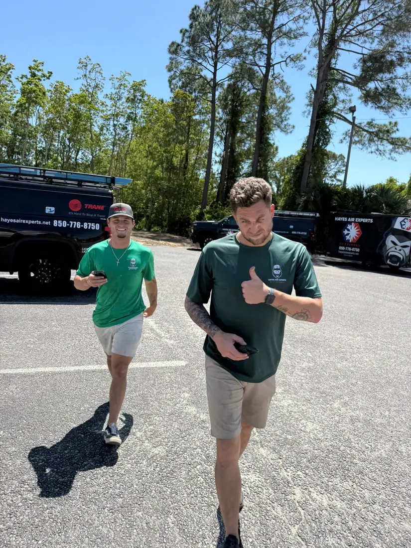 Two HVAC technicians in green Hayes Air Experts shirts walking in a sunny parking lot with branded service vans