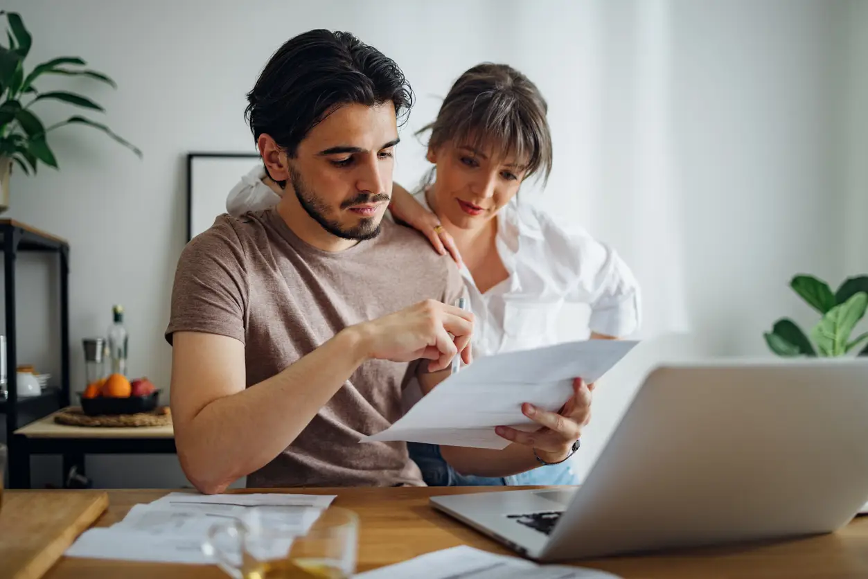 A man and woman reviewing documents at a laptop, discussing bills and paperwork at a kitchen table