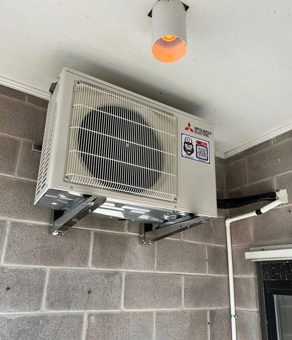 Mitsubishi Electric outdoor AC condenser unit wall-mounted on brackets under ceiling light against concrete block wall