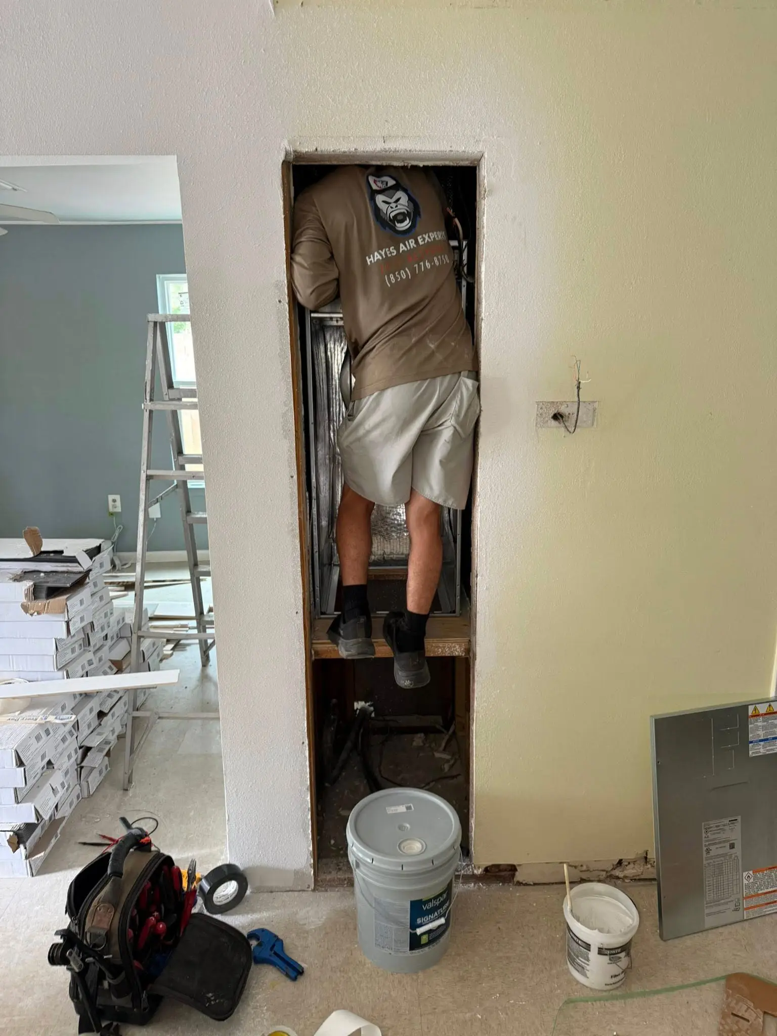 A worker climbing into a narrow wall cavity to service HVAC ductwork in a home under renovation, tools and ladder nearby.