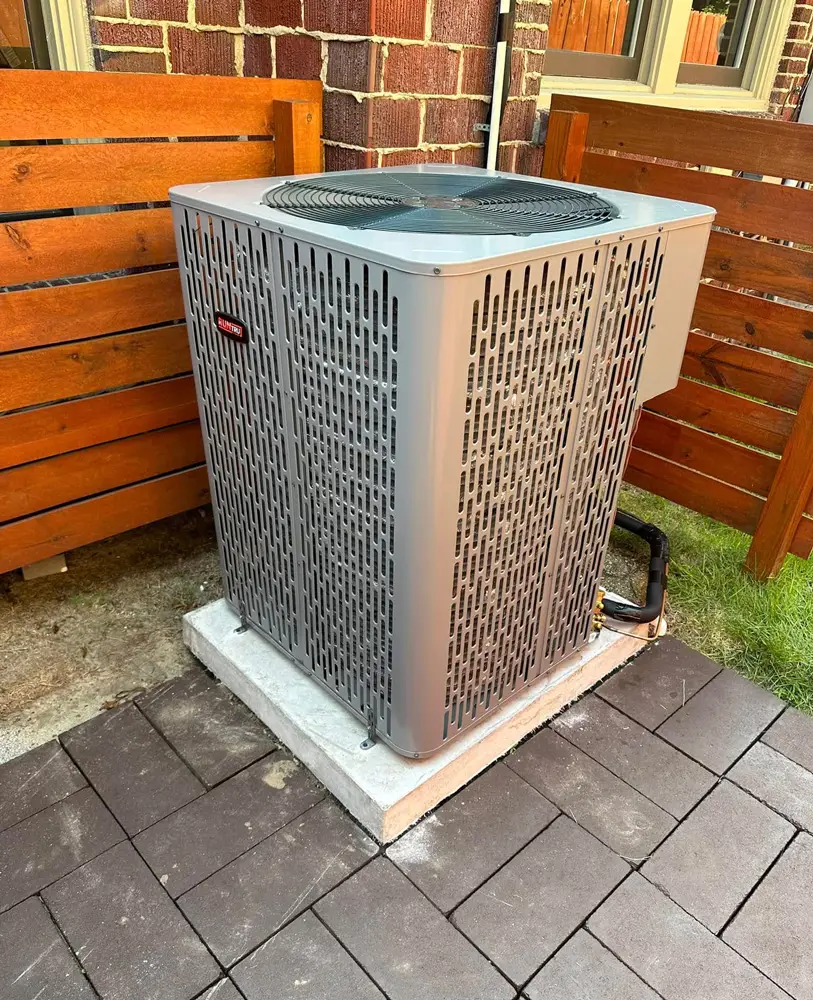 Outdoor HVAC condenser unit on concrete pad beside wooden fence and brick exterior