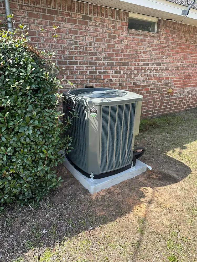 Outdoor central air conditioning unit on concrete pad beside brick house and shrubs
