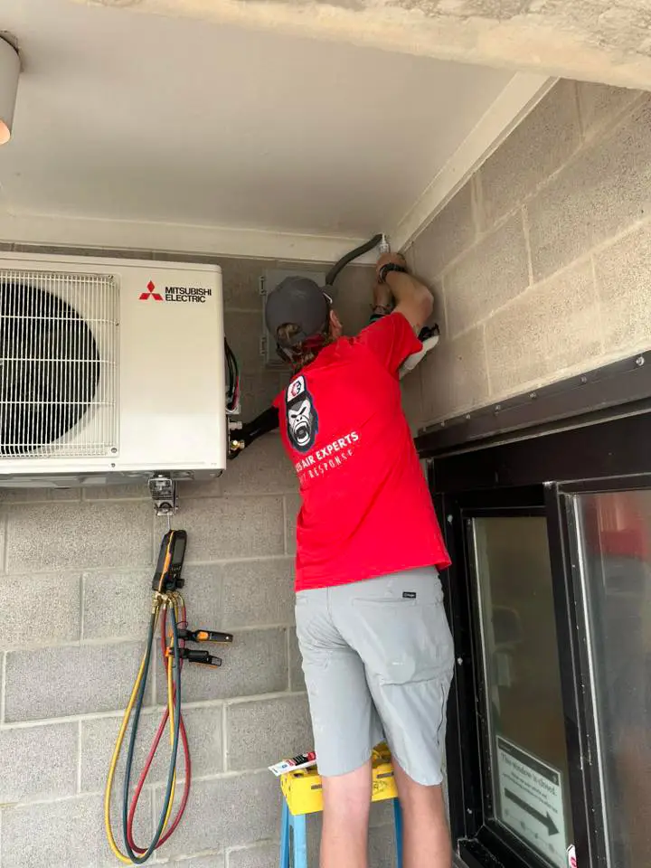 Technician in red shirt on ladder installing Mitsubishi Electric outdoor air conditioning unit on cinderblock wall