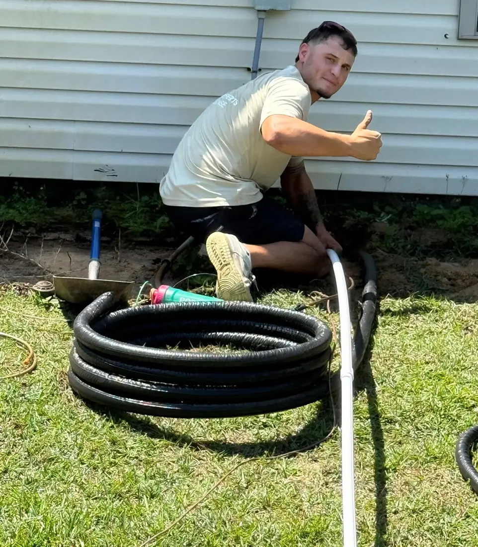 A man kneeling by house siding giving a thumbs-up while installing coiled drainage pipe with a shovel nearby.