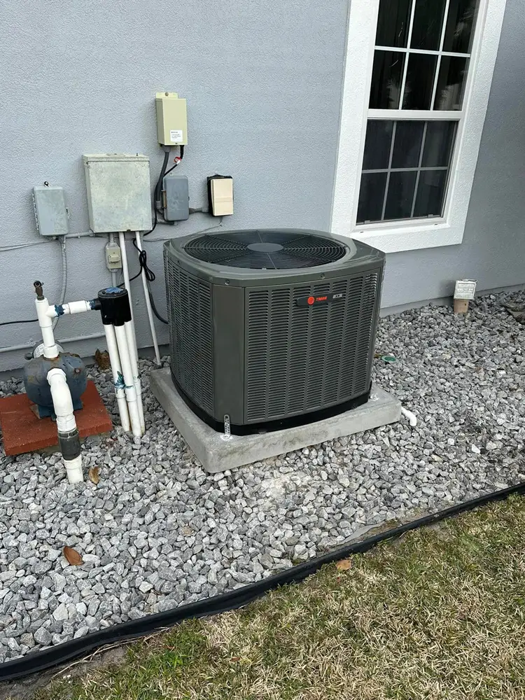 Trane outdoor AC condenser on concrete pad beside gray house, gravel bed, utility boxes and nearby window.