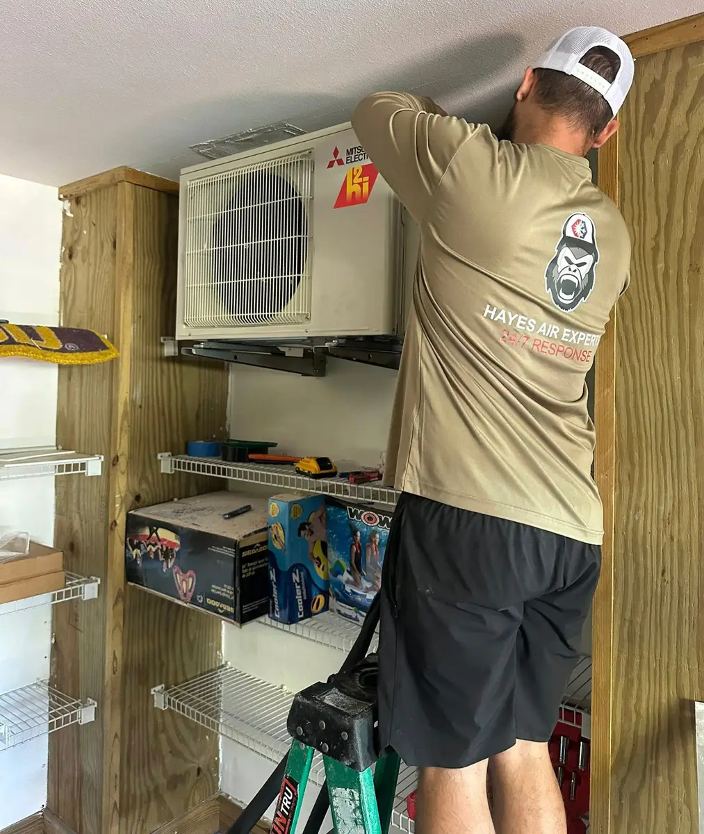 A man on a ladder installing a wall-mounted Mitsubishi mini-split HVAC unit inside a storage closet.