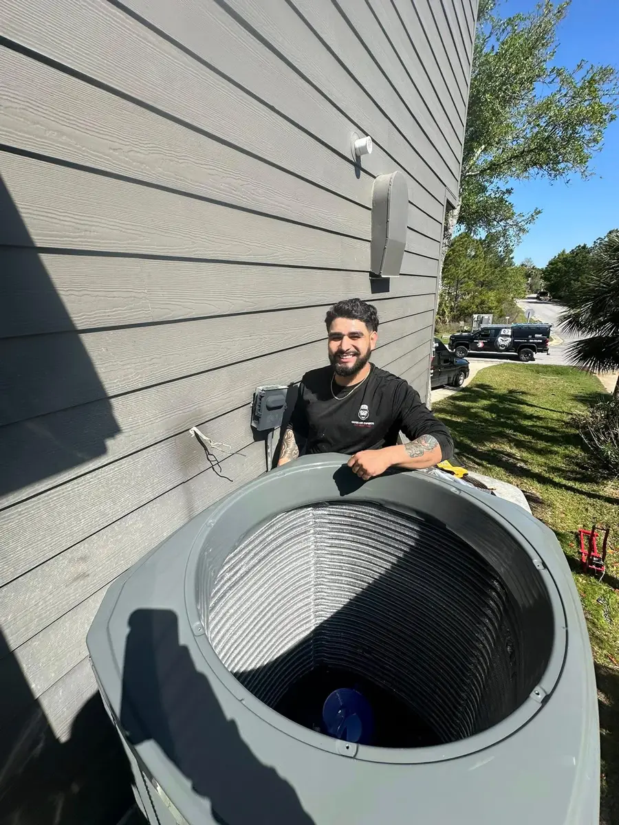A man HVAC tech inspecting an outdoor air conditioning condenser by house siding, work truck parked on the street.