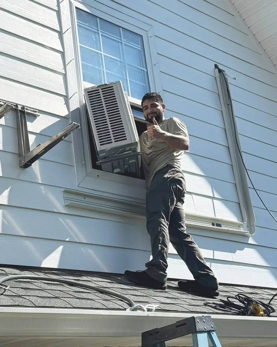 Man installing a window air conditioner, standing on a roof beside an open second-story window giving thumbs up.