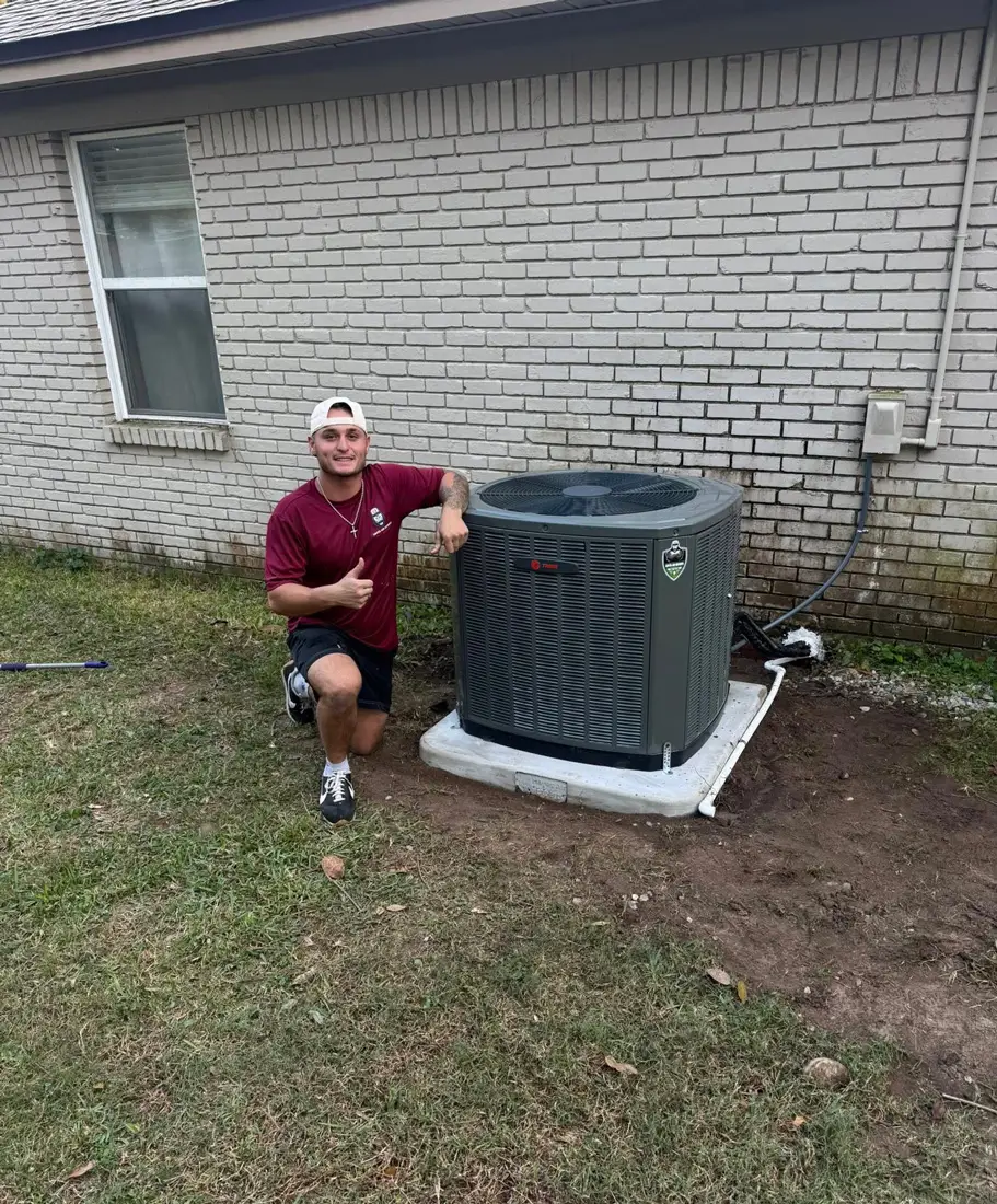 A man kneels beside a newly installed outdoor air conditioning unit next to a brick house, giving a thumbs up.